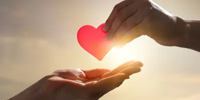 A hand holding a red paper heart above another outstretched hand, with the sun and sky in the background