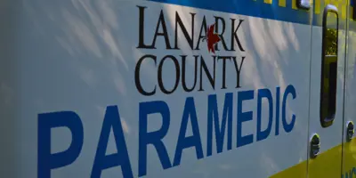 Close up the side of a white ambulance with blue and yellow decals along the top and bottom. Lettering on across the ambulance says "Lanark County Paramedic"